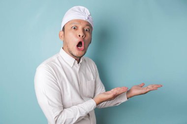 Shocked Balinese man wearing udeng or traditional headband and white shirt pointing at the copy space beside him, isolated by blue background