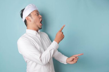 Shocked Balinese man wearing udeng or traditional headband and white shirt pointing at the copy space beside him, isolated by blue background
