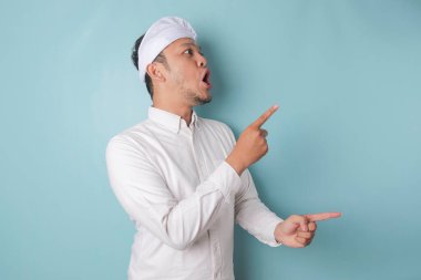 Shocked Balinese man wearing udeng or traditional headband and white shirt pointing at the copy space beside him, isolated by blue background