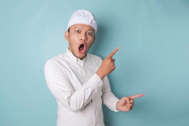 Shocked Balinese man wearing udeng or traditional headband and white shirt pointing at the copy space beside him, isolated by blue background