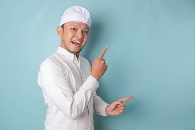 Excited Balinese man wearing udeng or traditional headband and white shirt pointing at the copy space beside him, isolated by blue background