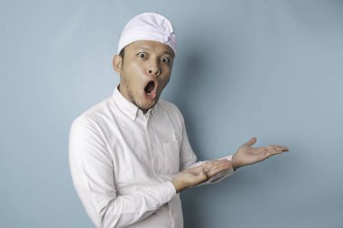 Shocked Balinese man wearing udeng or traditional headband and white shirt pointing at the copy space beside him, isolated by blue background