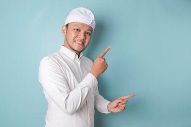 Excited Balinese man wearing udeng or traditional headband and white shirt pointing at the copy space beside him, isolated by blue background