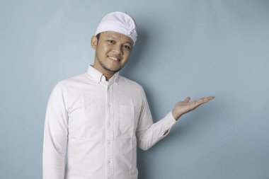 Excited Balinese man wearing udeng or traditional headband and white shirt pointing at the copy space beside him, isolated by blue background