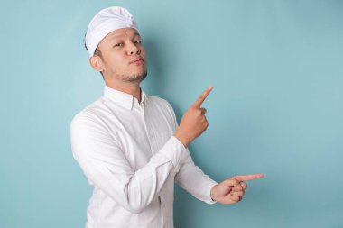 Excited Balinese man wearing udeng or traditional headband and white shirt pointing at the copy space beside him, isolated by blue background