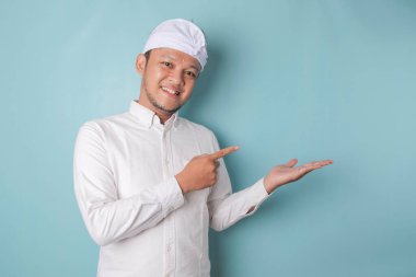 Excited Balinese man wearing udeng or traditional headband and white shirt pointing at the copy space beside him, isolated by blue background