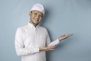 Excited Balinese man wearing udeng or traditional headband and white shirt pointing at the copy space beside him, isolated by blue background