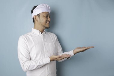 Excited Balinese man wearing udeng or traditional headband and white shirt pointing at the copy space beside him, isolated by blue background