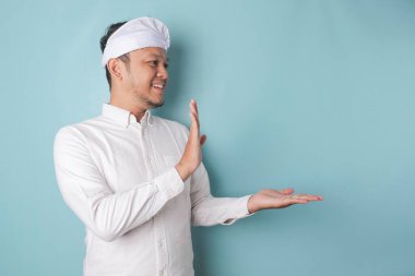 Excited Balinese man wearing udeng or traditional headband and white shirt pointing at the copy space beside him, isolated by blue background