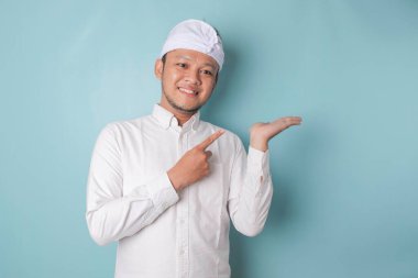 Excited Balinese man wearing udeng or traditional headband and white shirt pointing at the copy space beside him, isolated by blue background