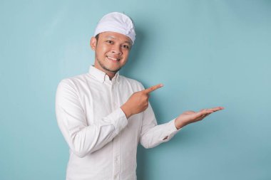 Excited Balinese man wearing udeng or traditional headband and white shirt pointing at the copy space beside him, isolated by blue background