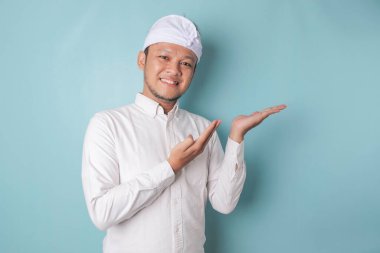 Excited Balinese man wearing udeng or traditional headband and white shirt pointing at the copy space beside him, isolated by blue background