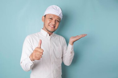 Excited Balinese man wearing udeng or traditional headband and white shirt gives thumbs up hand gesture of approval, isolated by blue background