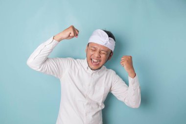 A young Balinese man with a happy successful expression wearing udeng or traditional headband and white shirt isolated by blue background