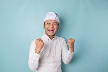A young Balinese man with a happy successful expression wearing udeng or traditional headband and white shirt isolated by blue background