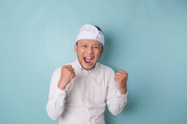 A young Balinese man with a happy successful expression wearing udeng or traditional headband and white shirt isolated by blue background