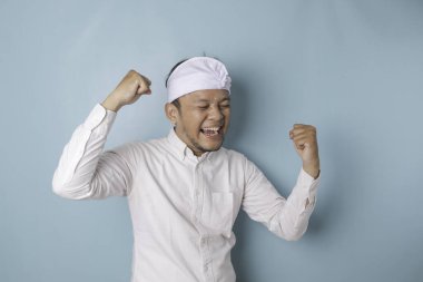 A young Balinese man with a happy successful expression wearing udeng or traditional headband and white shirt isolated by blue background