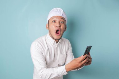 Surprised Balinese man wearing udeng or traditional headband and white shirt holding his smartphone, isolated by blue background
