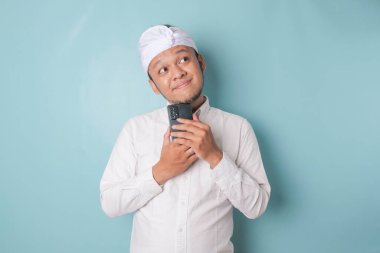 Portrait of a thoughtful young Balinese man wearing udeng or traditional headband and white shirt looking aside while holding smartphone isolated over blue background