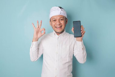 Excited Balinese man wearing udeng or traditional headband and white shirt gives OK hand gesture of approval while holding smartphone, isolated by blue background