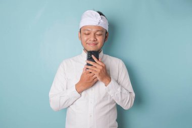 Portrait of a thoughtful young Balinese man wearing udeng or traditional headband and white shirt looking aside while holding smartphone isolated over blue background