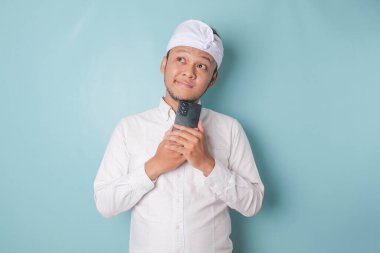 Portrait of a thoughtful young Balinese man wearing udeng or traditional headband and white shirt looking aside while holding smartphone isolated over blue background