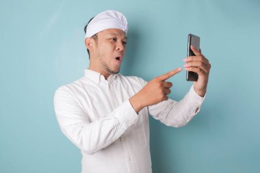 Surprised Balinese man wearing udeng or traditional headband and white shirt holding his smartphone, isolated by blue background