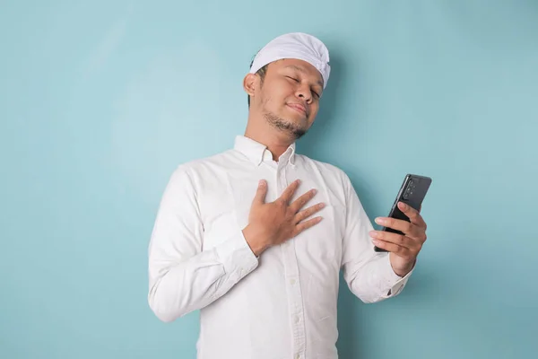 Happy mindful thankful young Balinese man holding phone and hand on chest smiling isolated on blue background feeling no stress, gratitude, mental health balance, peace of mind concept.