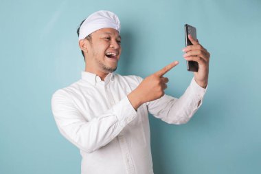 A portrait of a happy Balinese man is smiling and holding his smartphone wearing udeng or traditional headband and white shirt isolated by a blue background