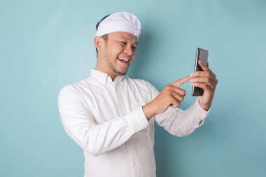 A portrait of a happy Balinese man is smiling and holding his smartphone wearing udeng or traditional headband and white shirt isolated by a blue background