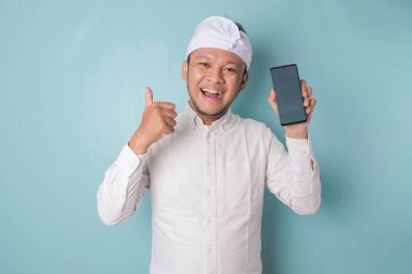 Excited Balinese man wearing udeng or traditional headband and white shirt gives thumbs up hand gesture of approval while holding smartphone, isolated by blue background