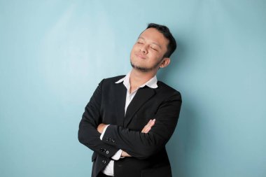 Portrait of a confident smiling Asian businessman standing with arms folded and looking at the camera isolated over blue background, wearing a black suit