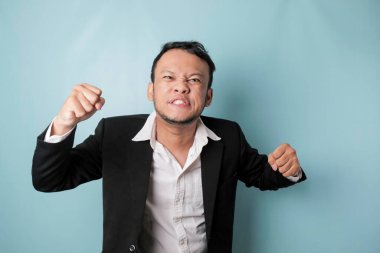 Angry and mad face of Asian businessman in black suit on isolated blue background.