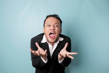 Angry and mad face of Asian businessman in black suit on isolated blue background.