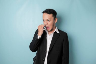 Asian man wearing black suit is picking his nose isolated on blue background.