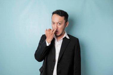 Asian man wearing black suit is picking his nose isolated on blue background.