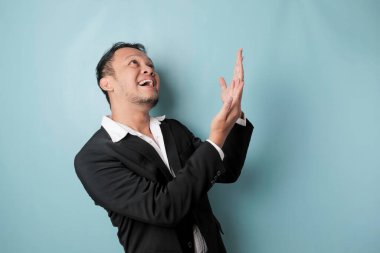 Excited Asian man wearing suit pointing at the copy space upside him, isolated by blue background