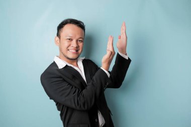 Excited Asian man wearing suit pointing at the copy space upside him, isolated by blue background