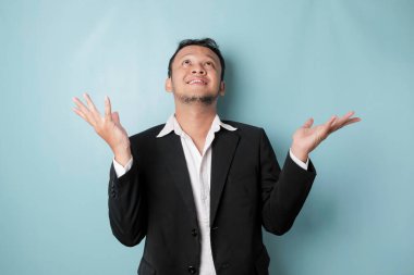 Excited Asian man wearing suit pointing at the copy space upside him, isolated by blue background