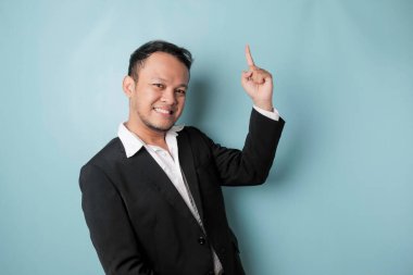 Excited Asian man wearing suit pointing at the copy space upside him, isolated by blue background