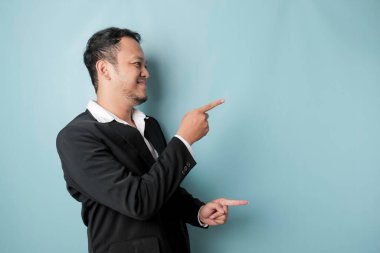 Excited Asian man wearing suit pointing at the copy space beside him and smiling, isolated by blue background