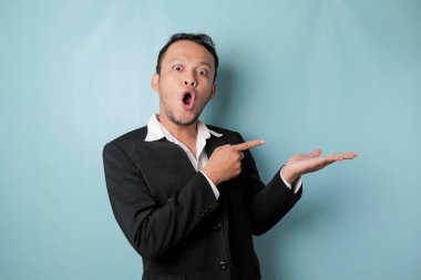 Shocked Asian man wearing suit pointing at the copy space beside him and smiling, isolated by blue background