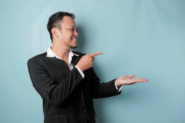 Excited Asian man wearing suit pointing at the copy space beside him and smiling, isolated by blue background