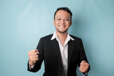 A young Asian man with a happy successful expression wearing suit isolated by blue background