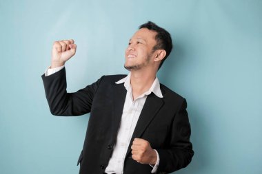 A young Asian man with a happy successful expression wearing suit isolated by blue background