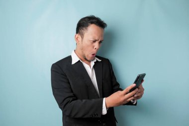 Surprised Asian businessman wearing black suit holding his smartphone, isolated by blue background
