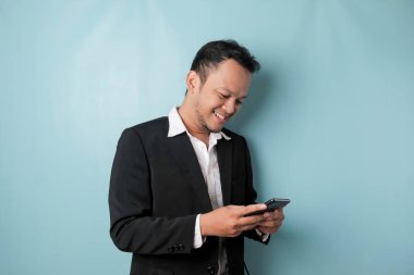 A portrait of a happy Asian businessman is smiling and holding his smartphone wearing black suit isolated by a blue background