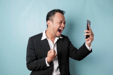 A young Asian man with a happy successful expression wearing suit and holding smartphone isolated by blue background