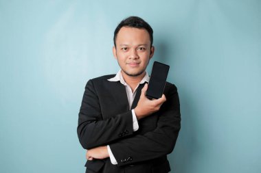 A portrait of a happy Asian businessman is smiling and holding his smartphone showing copy space on it's screen wearing black suit isolated by a blue background
