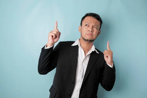 Shocked Asian man wearing suit pointing at the copy space upside him, isolated by blue background